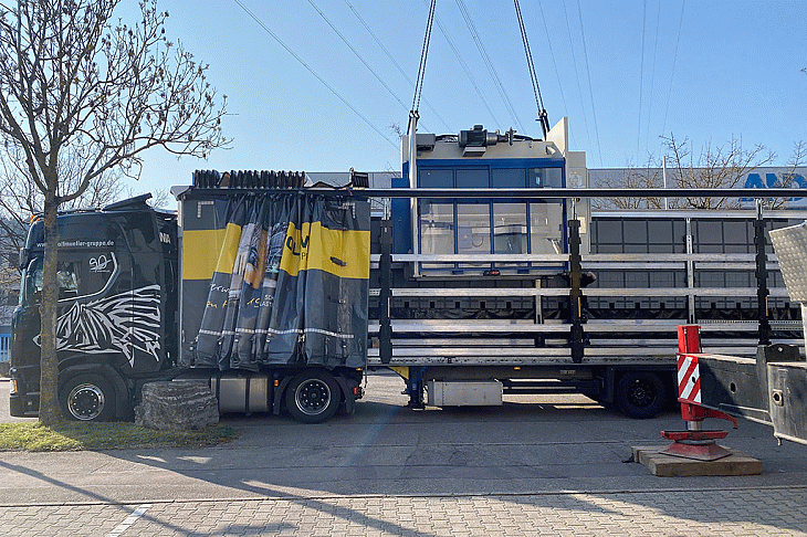 Eine große Maschine wird über das Dach des Sattelzuges  mit einem Kran auf den LKW gehoben